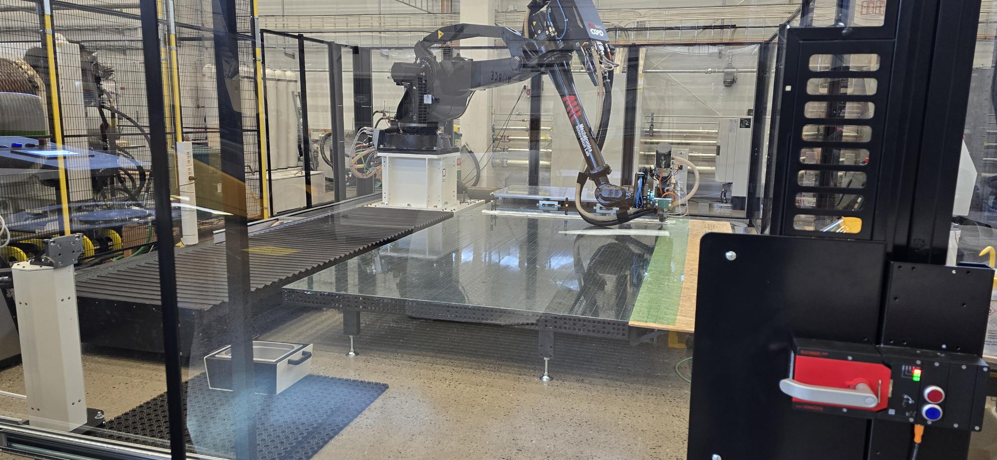 Printcellen hos Mechatronics Innovation Lab, sett gjennom sikkerhetsglasset. En robotarm med printhode for storformat-3D-print står over et stort byggebord inne i den inngjerdede cellen.